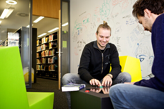 
                                Zwei Studierende sitzen sich gegenüber auf bunten Sitzgelegenheiten und spielen ein Computerspiel, das zwischen ihnen steht. 
                            